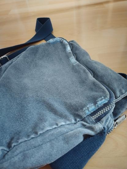 A dark blue, washed out small messenger bag on a wooden floor. The image shows the back of the bag, where woven mended patches in shades of blue and white stretch across the edges of the bag.