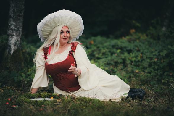 A person dressed in a fantasy-inspired costume resembling a mushroom sits on the forest floor. They wear a large, white, mushroom-shaped hat with lace trim, a cream-colored flowing dress with a red bodice, and have long, platinum blonde hair. Their makeup includes red and pink accents around the eyes. They hold a small red-and-white toadstool in one hand while gazing to the side with a playful expression. The setting is a lush, green forest with moss, small mushrooms, and foliage surrounding them.