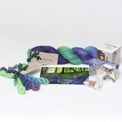 Picture shows a skein of yarn in shades of purple, blue and green. In front of the yarn is a photography of some bluebell woods. To the left is a mini skein bundle of yarn in shades of purple, blue and green. To the left is a box of tea, a bluebell soap and a white organza bag filled with buttons.
