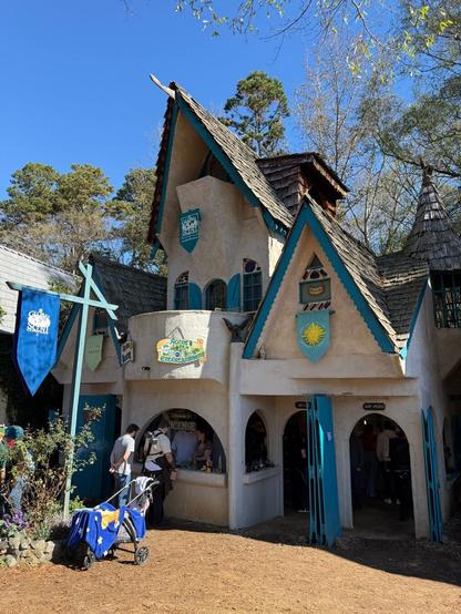 A whimsical, fairy-tale-style shop with steep roofs, blue shutters, and banners, selling incense and trinkets at a Renaissance fair.