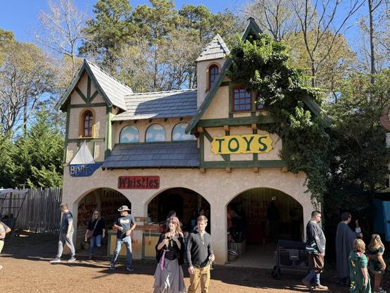 A medieval-style toy shop at a Renaissance fair, with ivy-covered walls, selling boats, whistles, and toys. People in costumes browse.