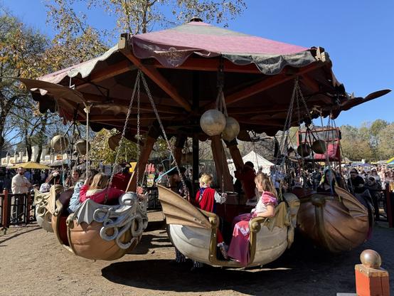 A whimsical, medieval-style carousel at a Renaissance fair, featuring dragon-themed swing seats under a red canopy, with costumed riders.