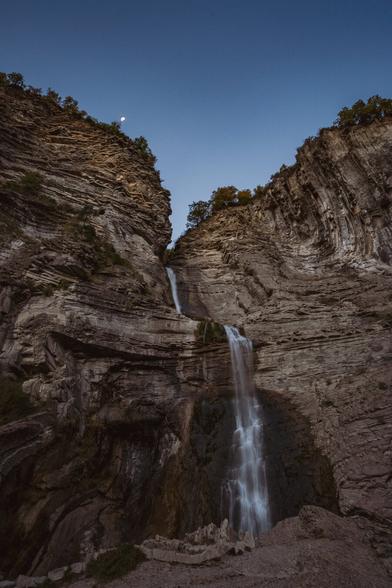 Vertical photograph showing a large steep stone wall from which a waterfall falls.
