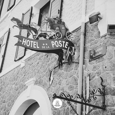 Black and white photograph capturing a street detail in Piriac-sur-Mer. The image focuses on a wrought iron hotel sign, featuring a stagecoach and bearing the inscription "Hôtel de la Poste". Right next to it, attached to the same building façade, a street plaque reads "Avenue A. Daudet", in tribute to the writer Alphonse Daudet.