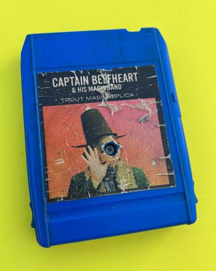 A photo of a vintage blue plastic 8-track cartridge tape showing excessive wear on a yellow background. It is entitled "Trout Mask Replica" by Captain Beefheart & The Magic Band. Inset photo is of an oddly dressed man wearing an old fashioned high-peaked black hat. He is holding something up in front of his face with his open-palmed right hand. Unfortunately the centre of the photo (his face) has been entirely obliterated by the screw holding the plastic tape frame together.