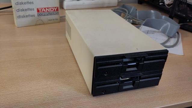 Two 5.25" floppy drives with a black front mounted on top of each other in a beige metal casing. A mains power lead and ribbon cable behind it are just out of focus.
