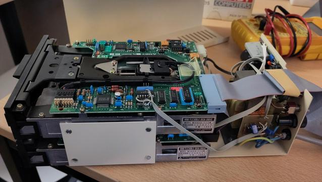 Two 5.25" disk drivers mounted on top of each other in a metal frame, the outer casing is removed revealing a green printed circuit board mounted on top of the top drive.