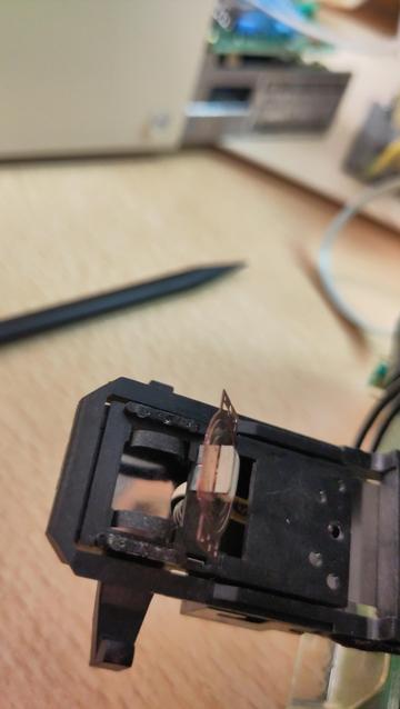 A black plastic rectangular part bent out of the casing of a floppy drive, the read/write head dangling from a flex pcb that is no longer attached to the plastic that is supposed to hold it.