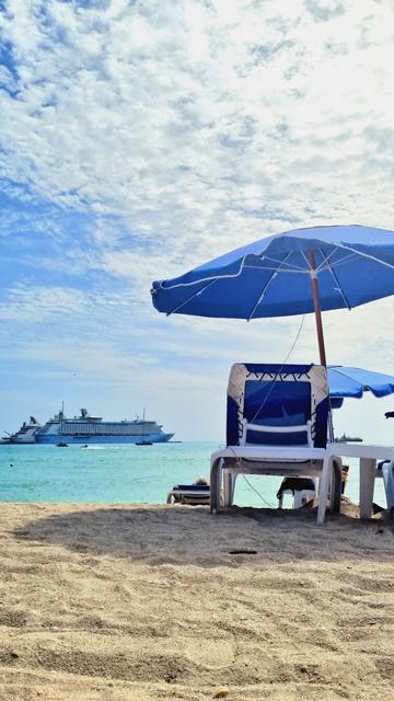 Blue sky, bright sun. White and blue beach chair and sun umbrella on the right. Golden sand of a beach next to the ocean and docked cruise ships in the distance.