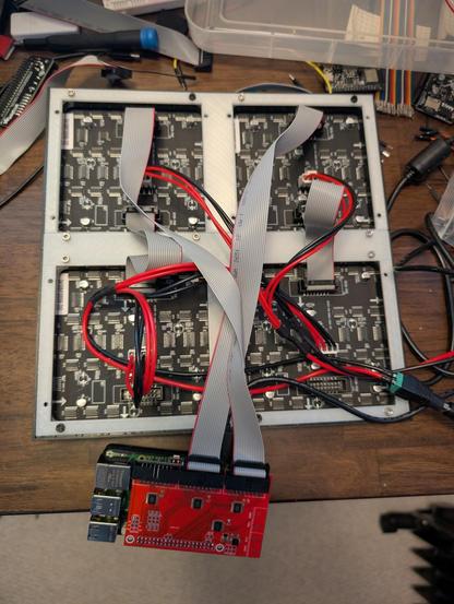 A view of the back of the matrix. You can see 4 individual sub panels held together by a white plastic 3D print. There are ribbon connectors and power cables, making the thing a bit of a rats nest. As in the other photo, it's on my messy desk with a dark wood finish & lots of unrelated wires, PCBs, tools & more.