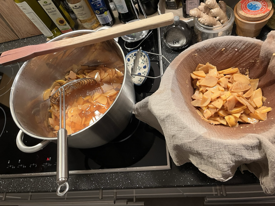 Ein Edelstahltopf mit Quittenschnitzen und Sud, in dem ein Schaumlöffel steht, daneben eine Schüssel mit einem Sieb, in dem ein Tuch mit Quittenschnitzen hängt; ein Kartoffelstampfer ist auch zu sehen