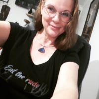 Blurry selfie of author L.E. Daniels, a fair-skinned woman with red-gold hair worn at shoulder length. She wears round glasses, a large pendant on a gold chain, and a black T-shirt that says "Eat the rude" in script. She smiles as if she has a secret, and her arms reach to the camera, as if they are holding it...or maybe she's coming out of your screen. Hard to be sure.