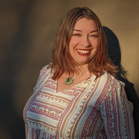 Color photo of Rebecca Gomez Farrell, a woman with brown eyes and light auburn hair. She wears a necklace with a flower pendant and a shirt of Mexican woven fabric. She stands in a sunbeam and against a tan background. She smiles warmly at the camera.