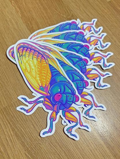 Photo of a small stack of periodical cicada stickers fanned out. The cicada is colored turquoise and pink and yellow instead of the actual insect's black and orange. It looks snazzy