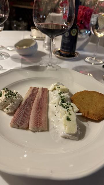Un plato gourmet con varias preparaciones de pescado (anguila y arenque). Incluye dos tiras de pescado ahumado, una ensalada cremosa con cebolla y hierbas frescas, una tostada crujiente y una porción de pasta de pescado con cebollino. El plato está servido en un elegante entorno de restaurante con copas de vino y decoración refinada en el fondo.