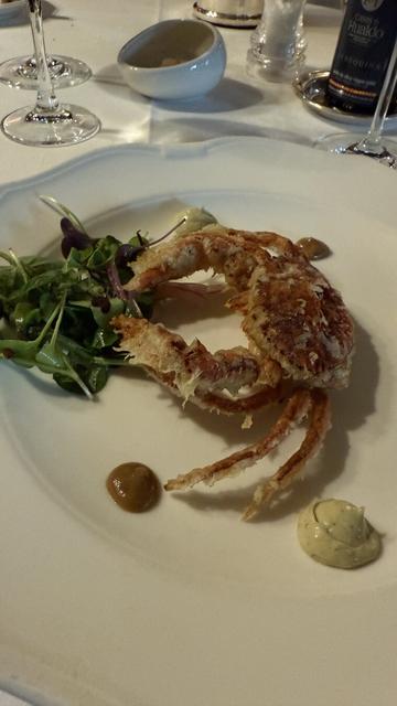 Un cangrejo de concha blanda frito, con una cobertura crujiente y dorada, servido con brotes verdes y salsas decoradas en puntos sobre el plato. La presentación es minimalista y refinada, en un plato blanco amplio, con copas de vino y una botella en segundo plano.