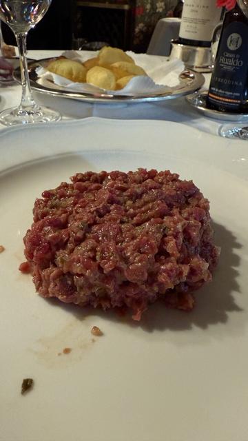 Un steak tartar de carne de ternera finamente picada y sazonada, servido en un plato blanco sin adornos. En el fondo, se observa un plato con patatas fritas crujientes, una botella de aceite de oliva y copas de vino, creando un ambiente de cena sofisticada.