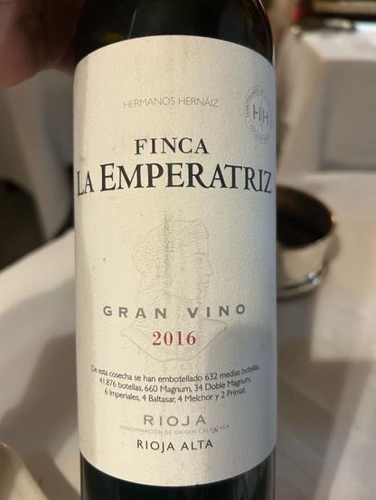 Una botella de vino tinto “Finca La Emperatriz Gran Vino 2016”, de la región de Rioja Alta, España. La etiqueta indica detalles sobre la producción y embotellado del vino. La botella es sostenida en una elegante mesa de restaurante, con un fondo desenfocado de copas y decoración.
