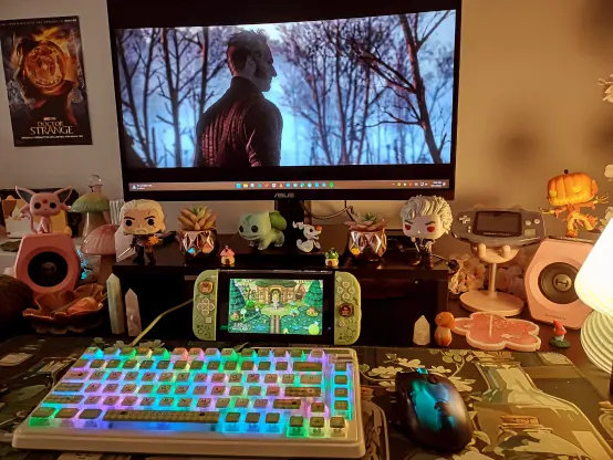 This is a photo of my gaming desk with sage and darker green shades, along with pops of pink. I have my Nintendo Switch with Animal Crossing's on the screen. It's sitting on my desk. I have Jack Skellington, Bulbasaur, Geralt, and Astarion Funko Pops beside my monitor, mushrooms everywhere, and crystals all around. Emiel Regis from The Witcher is my wallpaper on my computer.