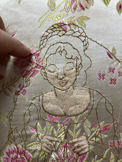 A close up view of a woman’s face outlined on a cream and pink rose brocade, slowly being filled with a pale skin tone in satin and long and short stitch.