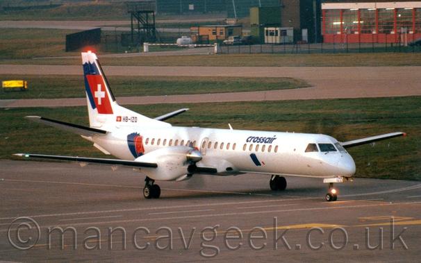 Side view of a white, twin propellor-engined airliner taxiing from left to right.
There are blue "CrossAir" titles on the upper forward fuselage, with blue and red splotches on the front and rear fuselage, as well as on the tail.
The red splotch on the tail is overlaid with a large white cross.
The registration "HB-IZO" is on the bottom of the tail in black, next to a Swiss flag, with the letters "IZO" also appearing on the open nose-wheel doors.
A black taxiway covered with white road markings and yellow arrows and lines fills the foreground, with more taxiways separated by large grassed areas fill most of the background, and a red, heavily glazed building at the edge of a compound surrounded by black chainlink fencing on the  upper left of the frame.