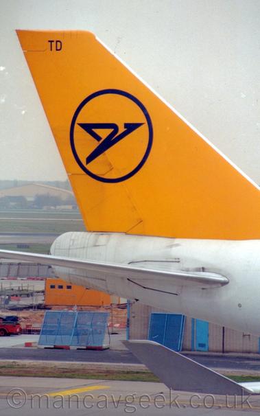 Closeup of the white rear fuselage and yellow tail of a very large jet airliner taxiing from left to right.
There is the blue outline of a circle on the tail, with a stylised blue flying bird in the middle.
The plane's white, upturned wingtip is visible in the bottom right corner of the framer.
Blue metal fences and a yellow pre-fabricated are visible under the plane's rear fuselage.
Behind the tail is a view across the airfield to a creamy-coloured hangar, under grey skies.