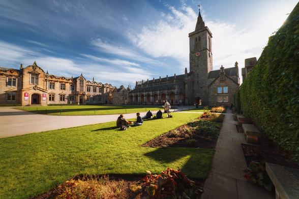 St Salvator's Quadrangle at the University of St Andrews, featuring a picturesque scene with historic stone buildings and a tall clock tower. The quadrangle is filled with lush green grass, where a group of people are seated, enjoying the outdoors. The buildings display intricate architectural details, including large windows and decorative elements, with some areas adorned with pink banners. In the foreground, a neatly paved path leads through the well-maintained gardens, lined with flower beds. Above, the sky is bright with scattered clouds, enhancing the inviting atmosphere of the university setting.