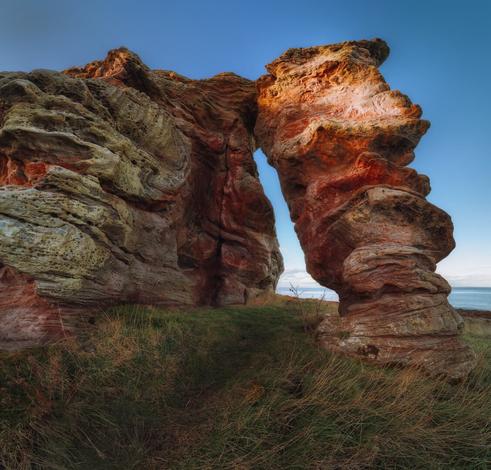 Buddo Rock at Buddo Ness, featuring impressive rock formations with intricate textures and colours. The towering red and orange hues of the stone contrast against a clear blue sky. A prominent archway is visible, formed by two large, weathered rock pillars that rise dramatically from the grassy ground. The surrounding area is dotted with tufts of grass, enhancing the natural setting, while the distant coastline can be glimpsed beyond the rock formations, adding to the scene's rugged beauty.