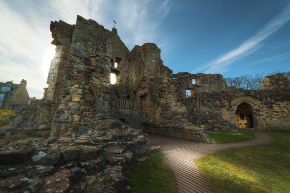 The ruins of St Andrews Castle, showcasing a weathered stone structure with remnants of walls and openings. The castle's stones are unevenly placed, with moss and lichen visible on some surfaces, hinting at its age. In the background, a blue sky with wispy clouds adds a serene atmosphere. A winding path made of wooden slats leads through the grassy area in front of the castle, guiding the viewer's eye towards the entrance of the structure, which features an arched doorway.