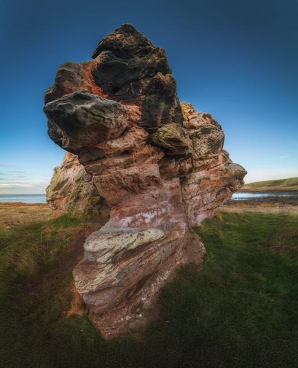 Another view of Buddo Rock at Buddo Ness, highlighting the unique geological formations of the area. The towering rock structure showcases an array of colours, including deep blacks, earthy reds, and soft greens, with intricate textures that suggest years of erosion. The sunlight casts gentle shadows, accentuating the contours of the rock. In the background, the coastline is visible, meeting the calm sea under a clear blue sky. The surrounding grass adds a natural element, contributing to the wild and rugged beauty of the landscape.