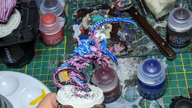A Tyranid painted in blue and purple. In the background are several paint pods and a green cutting mat.