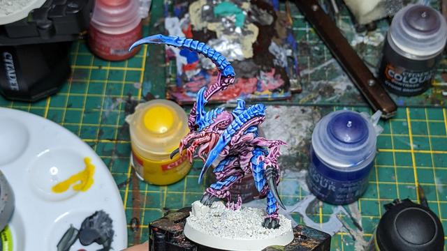 A Tyranid painted in blue and purple seen more from the front. In the background are several paint pods and a green cutting mat.