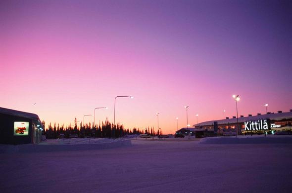 Bus station outside Kittilä airport. The airport building with the lettering lit is on the right, on the left a small building. In between and in the front the street and bus turn is empty, everything covered with thick crispy snow. Afar a row of dark pine trees. The whole picture is immersed in all shades of pink: The snow is dark lilac, the horizon above the trees glows in yellowish-pink, turning into reddish-pink upwards, and into blueish-lilac towards the right upper corner.