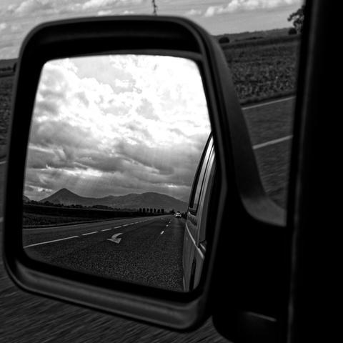 Vue depuis un rétroviseur de voiture : une route droite avec quelques voitures au loin, des montagnes à l’horizon et un ciel couvert de nuages. Photo en noir et blanc.