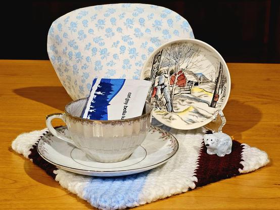 Tea tableau in white, blue, and purple with a vintage Johnson Brothers dish showing a winter farm scene, and antique opalescent white china, a cozy tea cozy, a tiny glass polar bear figurine, upon a striped, knitted square. The tea is Earl Grey Bella Luna loose leaf black tea from Adagio.