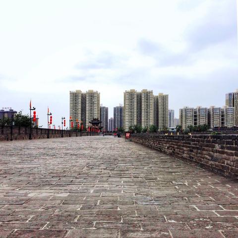 Walkway on top of the city wall with residential highrises on the horizon, red lanterns to the left, under a drizzly overcast sky. No people, except for small group in the distance.