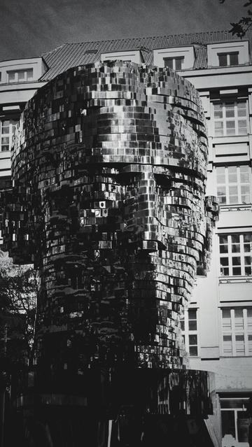 The image is in black and white and shows a large, metallic sculpture of a head. The sculpture is constructed from many horizontally stacked, segments of irregular shape that have mirror-like properties. Due to reflection, the left side of the head note appears black. The head is facing forward and is positioned outdoors in front of a building.
The building behind the sculpture has multiple stories and many windows. The windows are rectangular and have multiple panes.