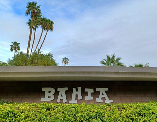Side of a low slung brown building with the word "BAHIA" in Polynesian multi-angled thick lettering. Palm trees and a blue sky partially obscured by clouds in the background. A hedge of green shrubs in the foreground.