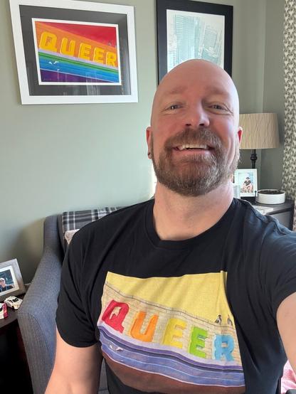 Me, bald and bearded, in a colorful tee that says QUEER in front of piece of art on my office wall by the same artist that says the same thing.