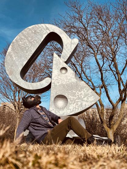 In this outdoor photograph, a figure reclines against a monumental metallic sculpture that dominates the composition with its bold geometric presence. The sculpture features a massive C-shaped form paired with a triangular cheese, both crafted from polished stainless steel that captures and reflects the brilliant blue sky overhead.

The human subject, wearing a distinctive black neoprene puppy hood that obscures their face while creating a striking silhouette, sits casually at the base of the structure. Their outfit consists of a textured gray jacket layered over a patterned button-up shirt, complemented by olive-brown pants and casual footwear. The juxtaposition of the figure's organic form against the sculpture's rigid geometry creates a compelling visual narrative—one where human and artistic expression intersect in unexpected ways.

Surrounding this central scene, dormant trees with bare branches reach toward the cloudless cerulean sky, their intricate network of limbs creating a natural frame that emphasizes the sculpture's artificial precision. The setting appears to be a public park or installation space during late winter or early spring, evidenced by the dried golden grasses carpeting the ground beneath the figure.

Light plays a crucial role in this composition, with the afternoon sun casting subtle shadows while simultaneously illuminating the sculpture's metallic surface, creating highlights that seem to shift and dance across its curves. This interplay of light, 