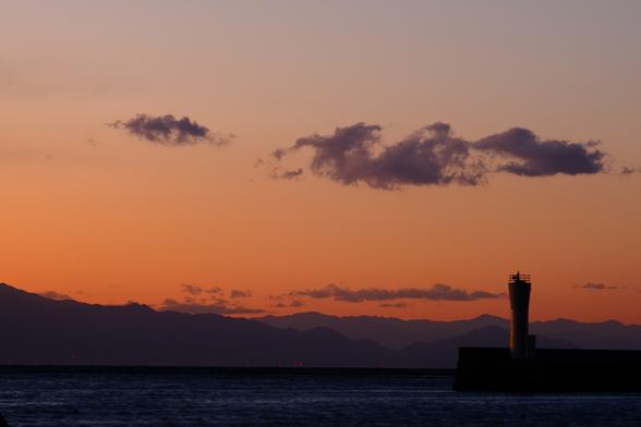 At the horizon, deep purple mountain foothills lie under an orange sky. In the foreground, a small lighthouse on an outcropping sits in a bay.
