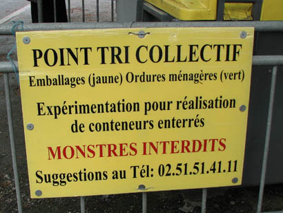Panneau jaune de déchetterie portant notamment les inscriptions :Point tri collectif / Emballages (jaune) Ordures ménagères (vert). Expérimentation pour réalisation DD conteneurs enterrés. Monstres interdits (en rouge)