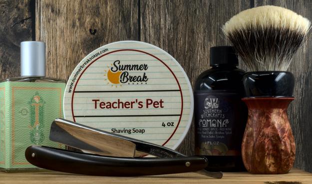 (Le Chat Mistral description)

This image shows grooming items arranged on a wooden surface. On the left, there's a green bottle with a geometric design. In the centre, a round tub labelled "Summer Break Soaps" features a "Teacher's Pet" shaving soap design with a notebook pattern. To the right, a dark bottle contains "Southern Witchcrafts Pomona" aftershave splash. In front, a straight razor with a dark handle lies horizontally. Beside it, a shaving brush with a wooden handle and white bristles stands upright. The scene has a rustic, masculine feel.