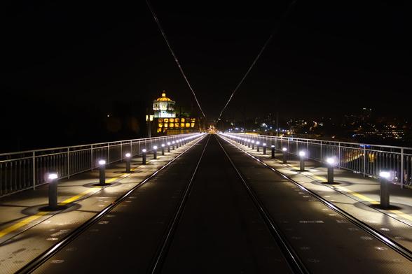A nighttime shot looking straight down the centre of a bridge, illuminated by evenly spaced, glowing bollard lights lining both sides. The bridge's surface is dark, with parallel tram tracks running along its length, disappearing into the distance. A building with lit windows and a domed tower is visible in the middle distance, framed by the bridge's structure and the dark night sky. The bridge's railings are visible on either side, and a faint cityscape with scattered lights is seen further in the background. Overhead wires, presumably for the trams, stretch diagonally upwards from the top centre of the frame, adding to the perspective and leading the eye towards the building.