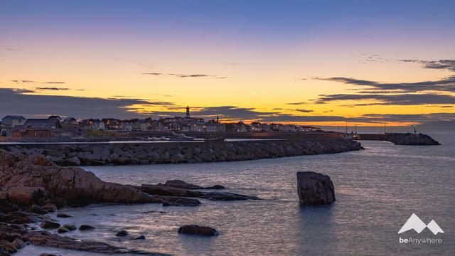 Sunset over a small fishing village on the coast. You can already see lights burning in the houses. The top of a lighthouse towers above it all.