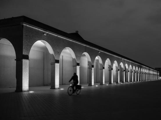 A black and white shot shows the seemingly endless row of arches with pillars of the "Venetian Galleries" in Ostend, under a dark sky. A person on a bicycle rides along the dark-colored brick path in front of the arches. The arches are illuminated from above and below, creating a stark contrast with the dark surroundings. The building above the arches has a simple, flat roofline. The perspective of the shot emphasizes the length of the arcade.