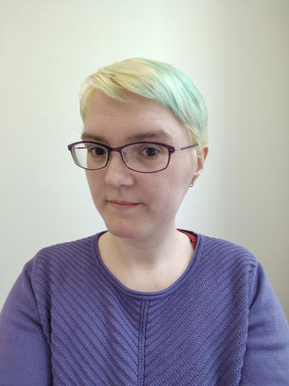 Selfie einer Frau mit Brille und kurzhaarschnitt in hell gebleichtem Blond mit ein paar türkisen Farbspitzen. Sie trägt einen Lila Pulli mit Strickmuster. Der Hintergrund ist eine weiße Wand.