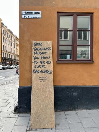 a board leaning against the orange-painted wall of a building. on it is written “your yoga class taught you to bend over backwards” and in smaller stencil lettering below that, “that doesn’t sound like fun to me”