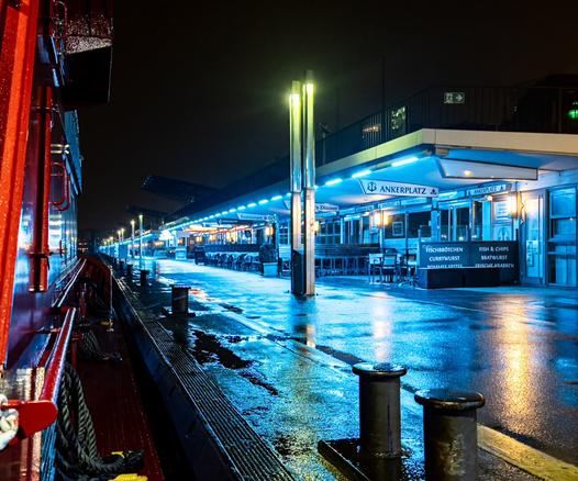 Die Bordwand einer Fähre die längsseits an einem Ponton liegt. Der Poton und die Gebäude auf dem regennassen Ponton sind bläulich beleuchtet. Alles unter nächtlichen Himmel.
