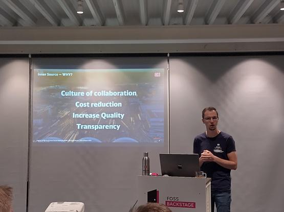 Carsten Hoffmann giving a talk standing in front of a slide that claims InnerSource brings a culture of collaboration, cost reduction, increased quality, transparency